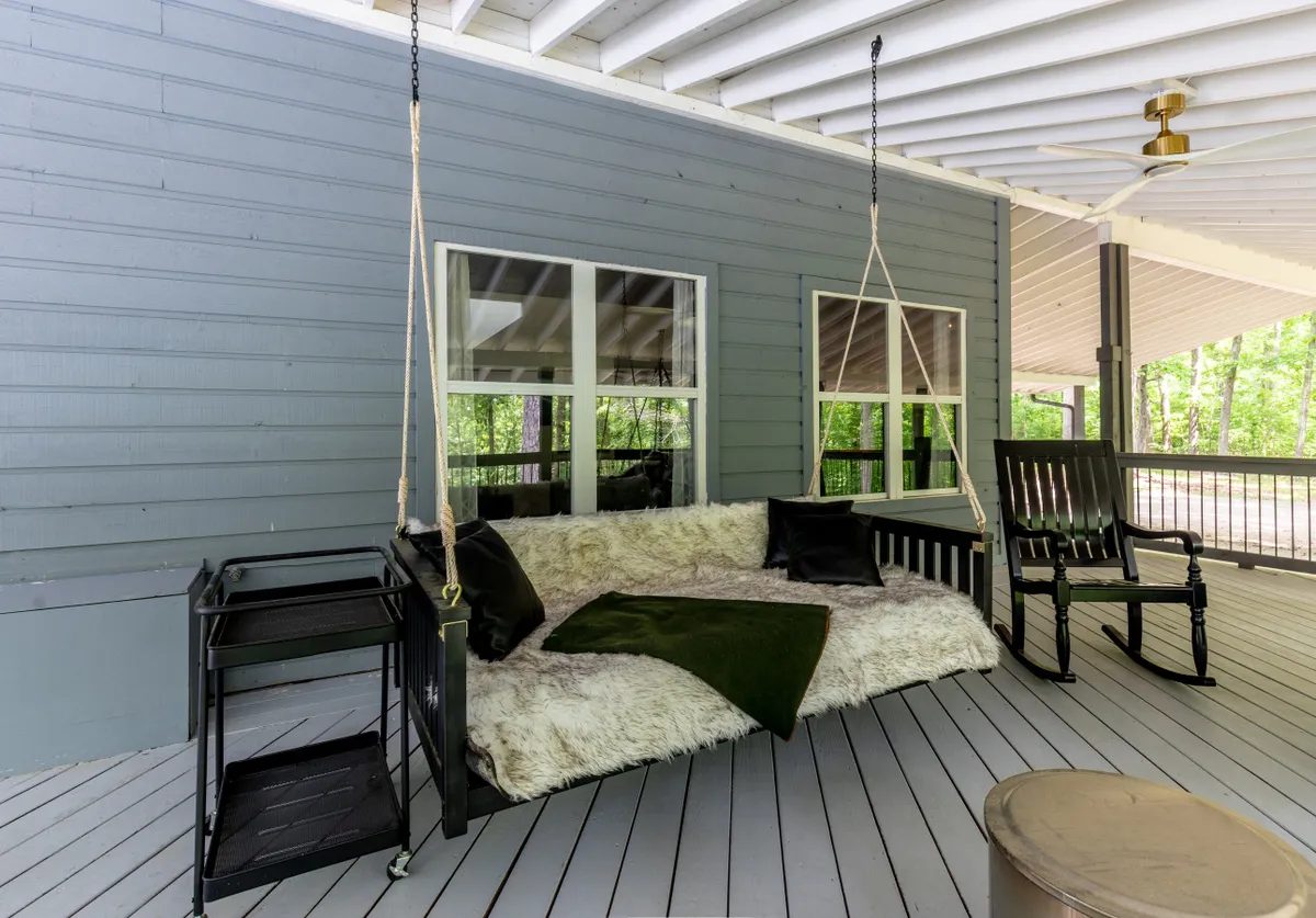 A tranquil covered front porch featuring a swinging couch, a rocking chair, and a black cart, all positioned to overlook the spacious yard and lush greenery. The area exudes comfort, with morning sun or evening breezes inviting guests to linger.