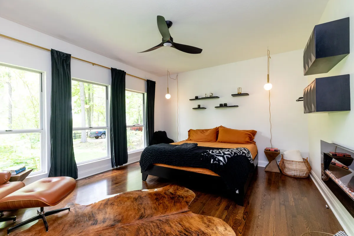 A contemporary bedroom featuring a bed with vibrant orange bedding, large windows with dark curtains framing views of nature, modern wall shelving, a sleek ceiling fan, and a comfortable leather lounge chair. The space is filled with natural light and styled for both rest and leisure.