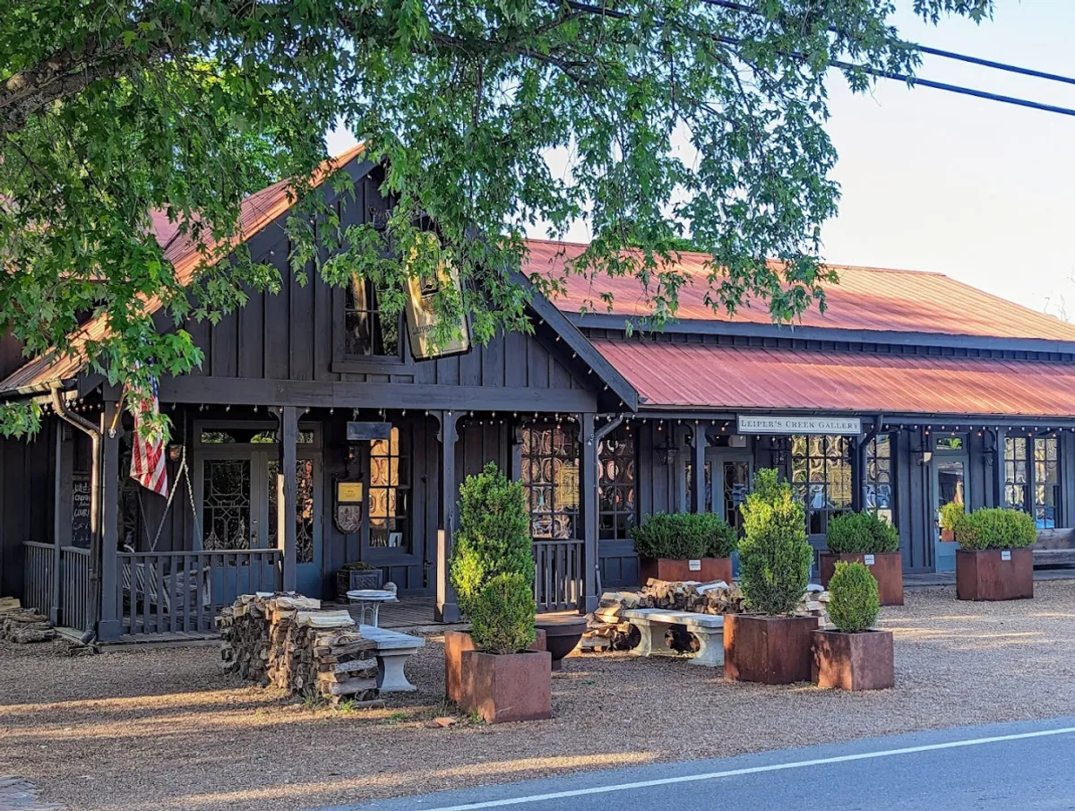 A distinctive exterior view of a main building with dark wood siding, a red metal roof, decorative windows, and planters overflowing with greenery. Logs are stacked at the side, adding rustic charm. The scene suggests a vibrant, welcoming environment—perfect for discovering local art or participating in creative community events.