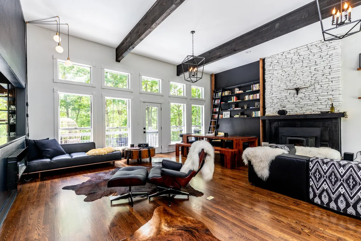 This image showcases an inviting living area with large windows, exposed wooden beams, a stone fireplace, a bookshelf filled with books, and stylish furniture. Sunlight pours into the space, highlighting the cozy seating area and the overall open, welcoming layout.