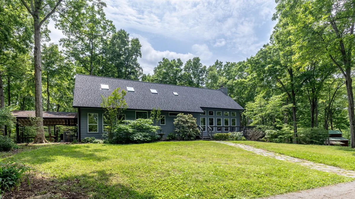The house is set among mature trees with a welcoming pathway leading up to the porch, evoking a tranquil escape. The image hints at ample outdoor space for gatherings and relaxing mornings with coffee.