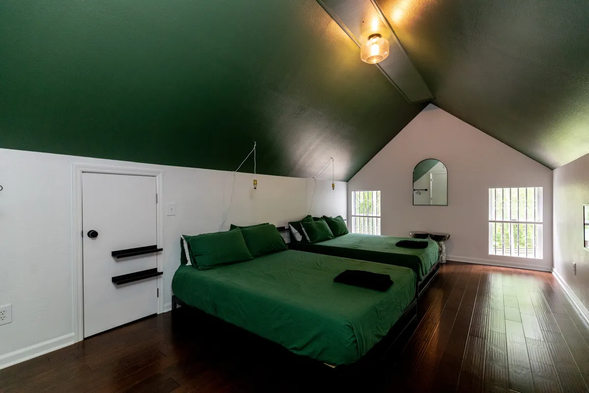 A spacious bedroom with two queen beds dressed in green bedding, a large wall mirror, and ample natural light streaming through big windows. The room exudes calm and is designed to accommodate families or groups.