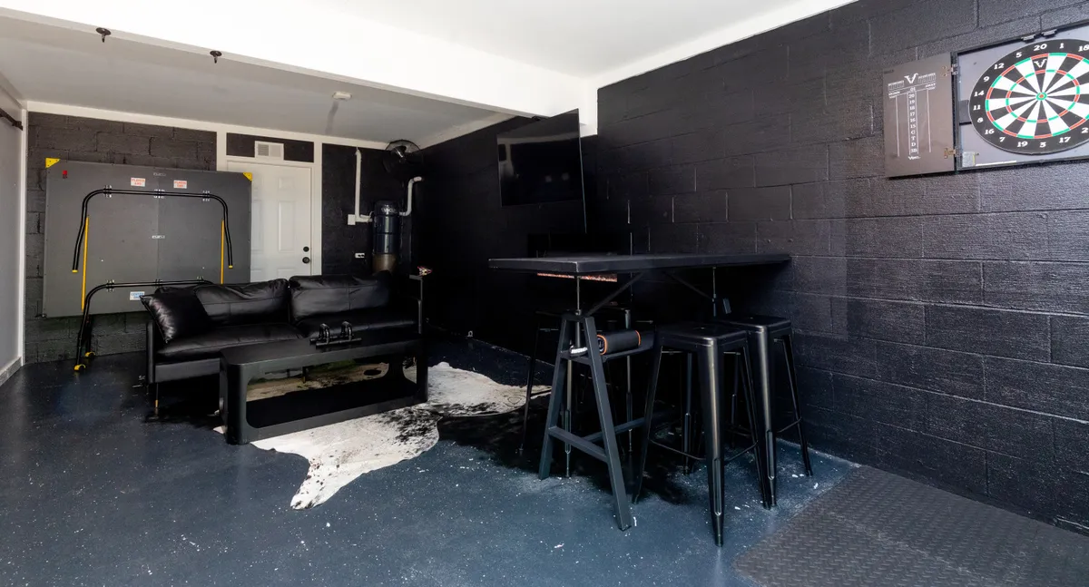 A relaxed basement recreation room featuring a black couch, a dartboard mounted on the wall, and a high-top table with stools. The space is styled for informal gatherings, with an emphasis on entertainment and game night activities.