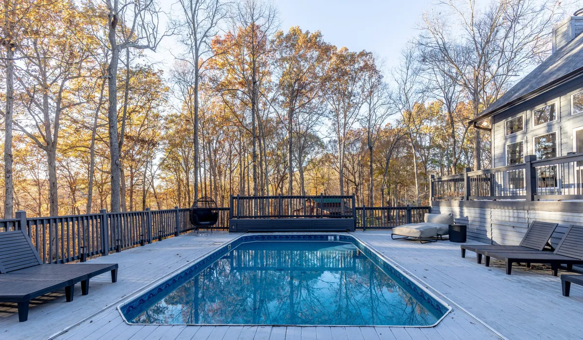 A serene in-ground swimming pool surrounded by tall trees and an adjacent deck area with outdoor seating. The scene is tranquil, ideal for sunbathing, swimming, or leisurely gatherings during the warmer months.