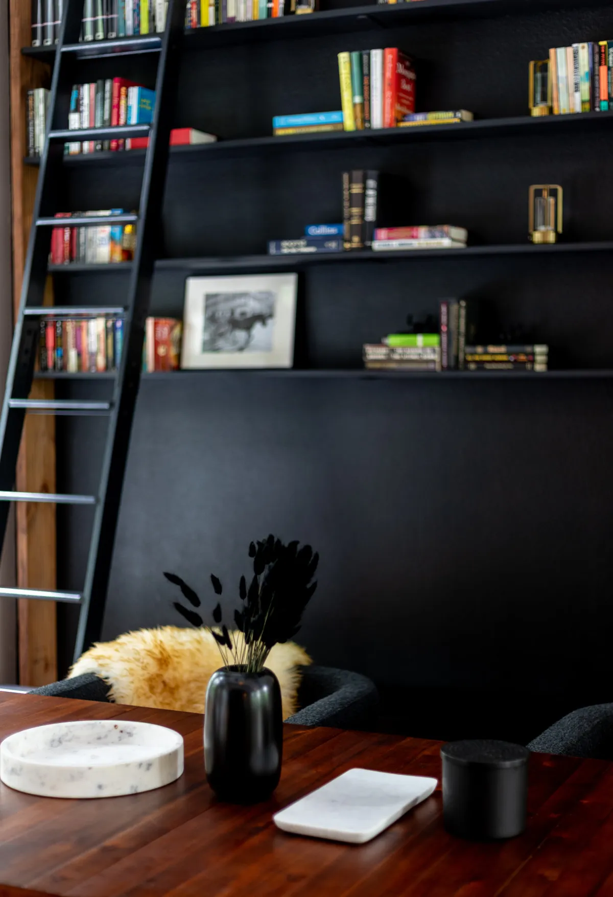 A bright loft space with bookshelves filled with books, a decorative ladder, a black vase with dried plants, and a wooden table. The setting is perfect for reading, writing, or quiet creative moments.