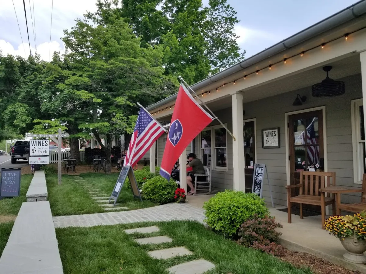 A lively outdoor patio featuring American and Tennessee flags, patio seating, and signage highlighting wine tastings and live music events. The garden-like setting encourages socializing and festive activities.