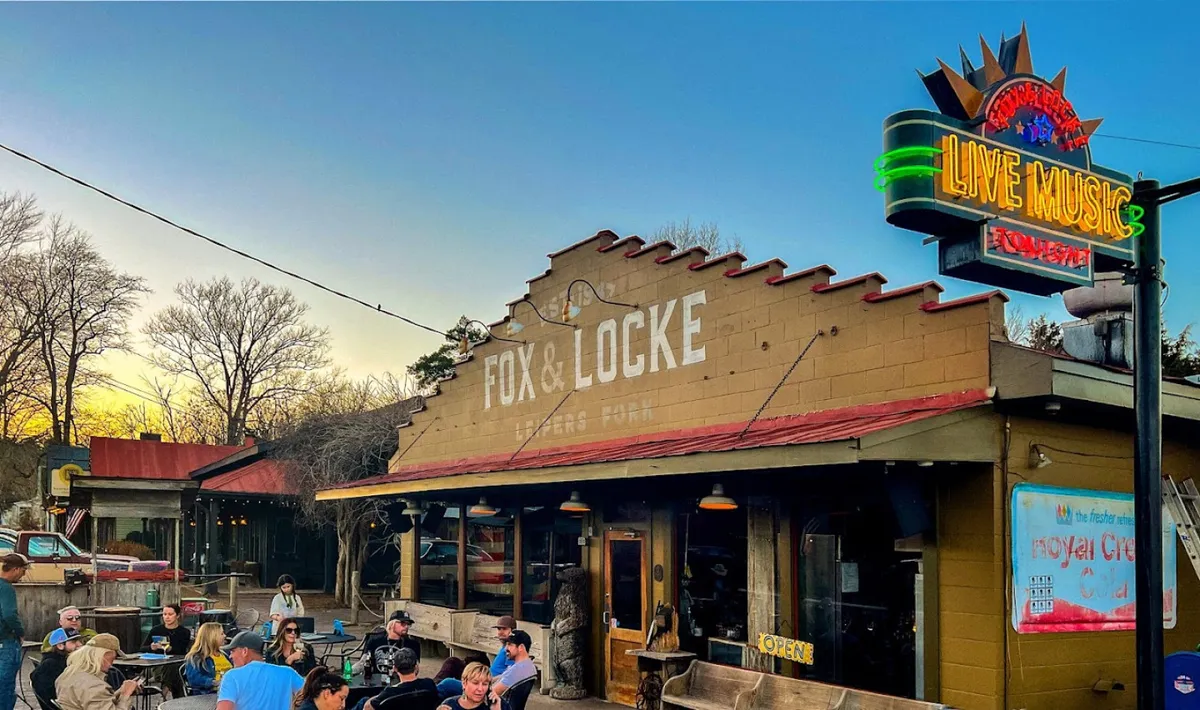 This image showcases the facade of a lively local restaurant featuring a neon sign, outdoor seating area, and a banner advertising live music. The scene exudes a festive, community atmosphere under the warm glow of sunset.